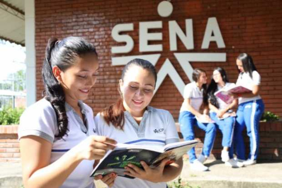 Carreras-del-SENA-mejor-pagadas Dos estudiantes del SENA mirando un cuaderno con una pared con el logo del SENA en el fondo.