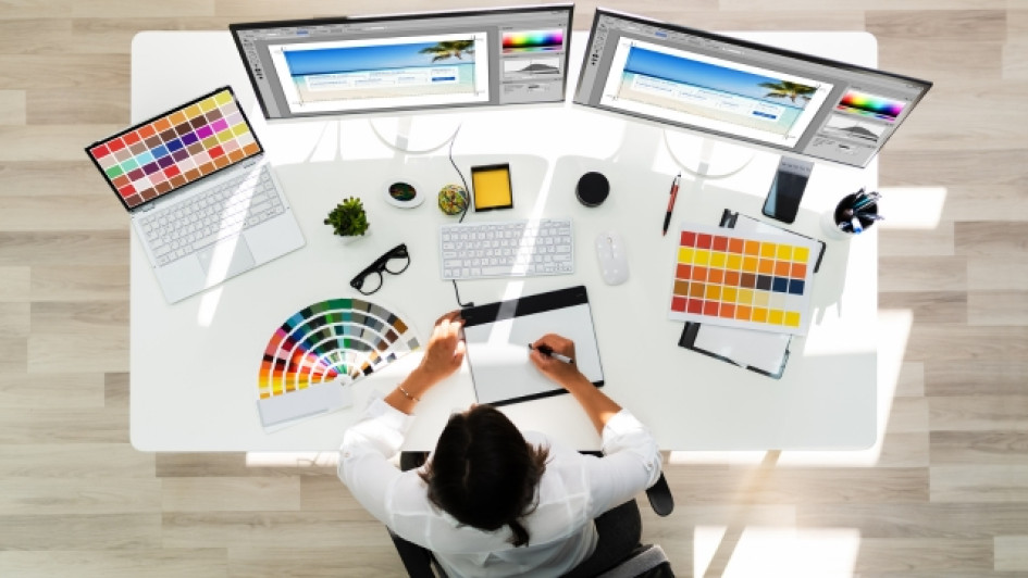 Cuanto-gana-un-diseñador-grafico-en-Colombia Vista superior de un diseñador gráfico trabajando en una mesa blanca con muchas pantallas, hojas y herramientas