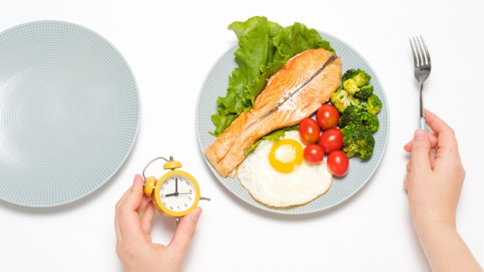Plan-de-alimentación-ayuno-intermitente Persona sentada frente a un plato de comida sosteniendo un reloj en su mano izquierda.